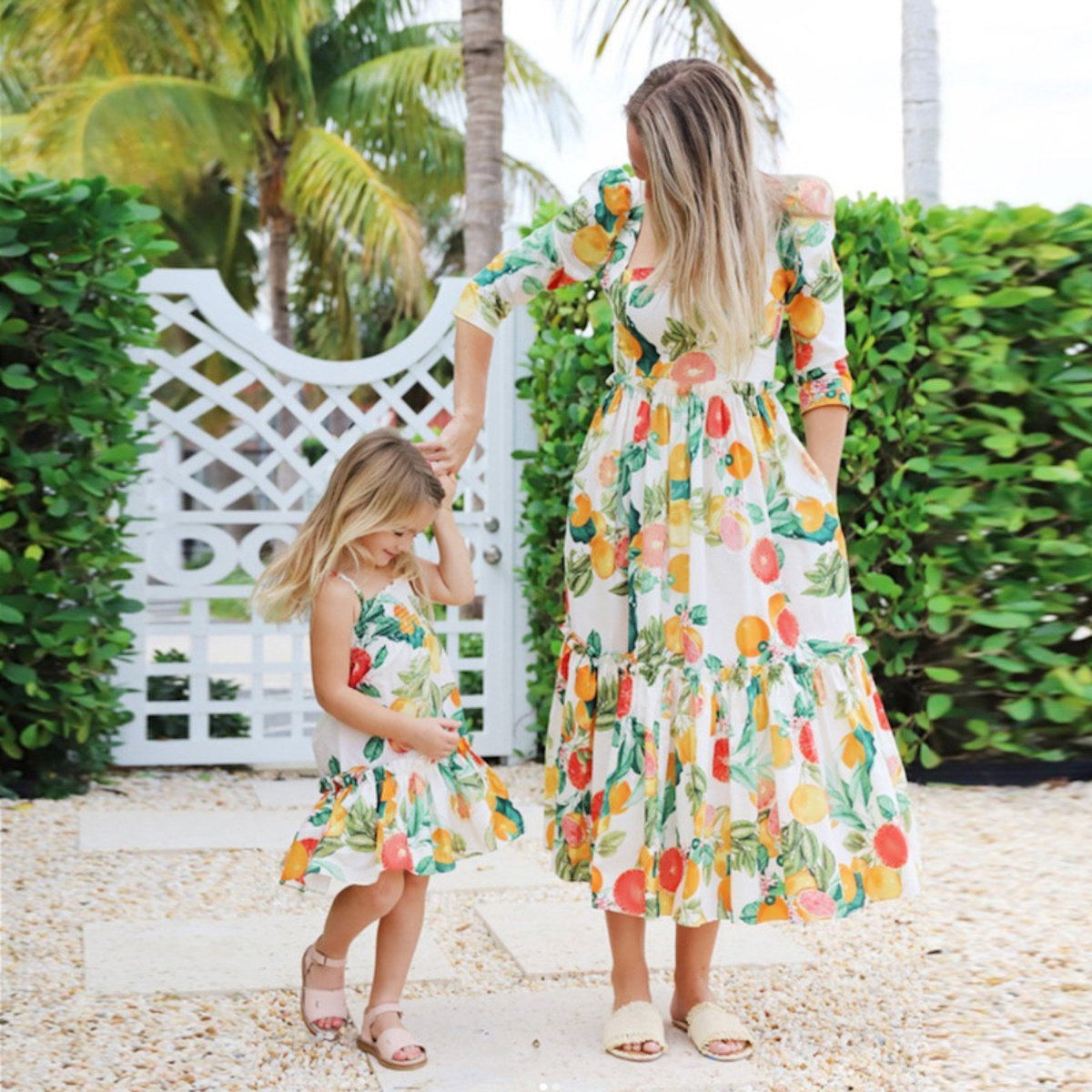 Two women in matching floral dresses standing outdoors with greenery and a white lattice structure in the background.