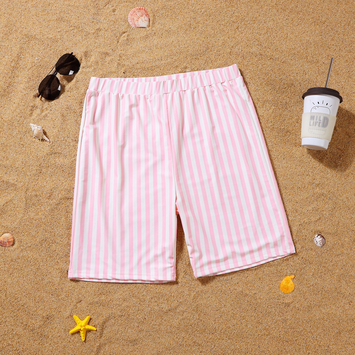 Pink and white striped shorts on a sandy surface with sunglasses and a cup.