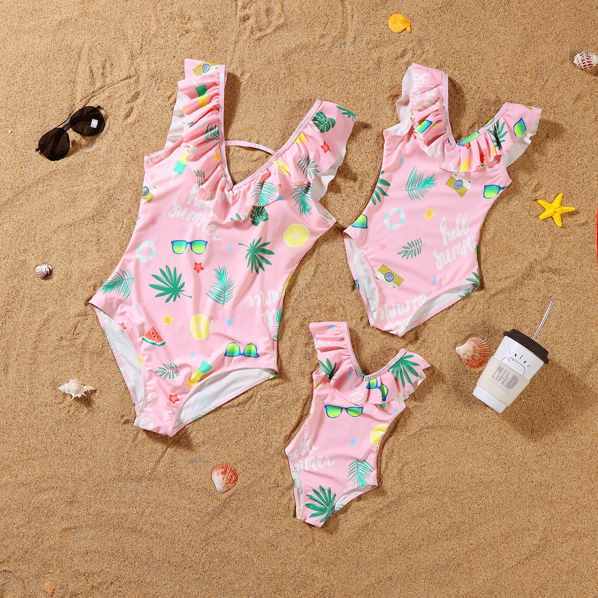 Three pink children's swimsuits with tropical patterns on a sandy surface.