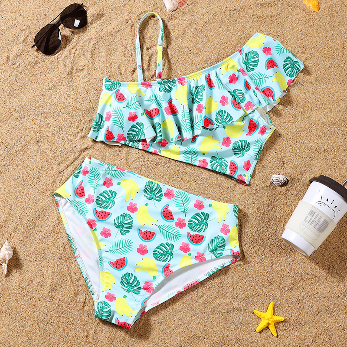 Two-piece swimsuit with tropical pattern on sand with sunglasses and sunscreen.